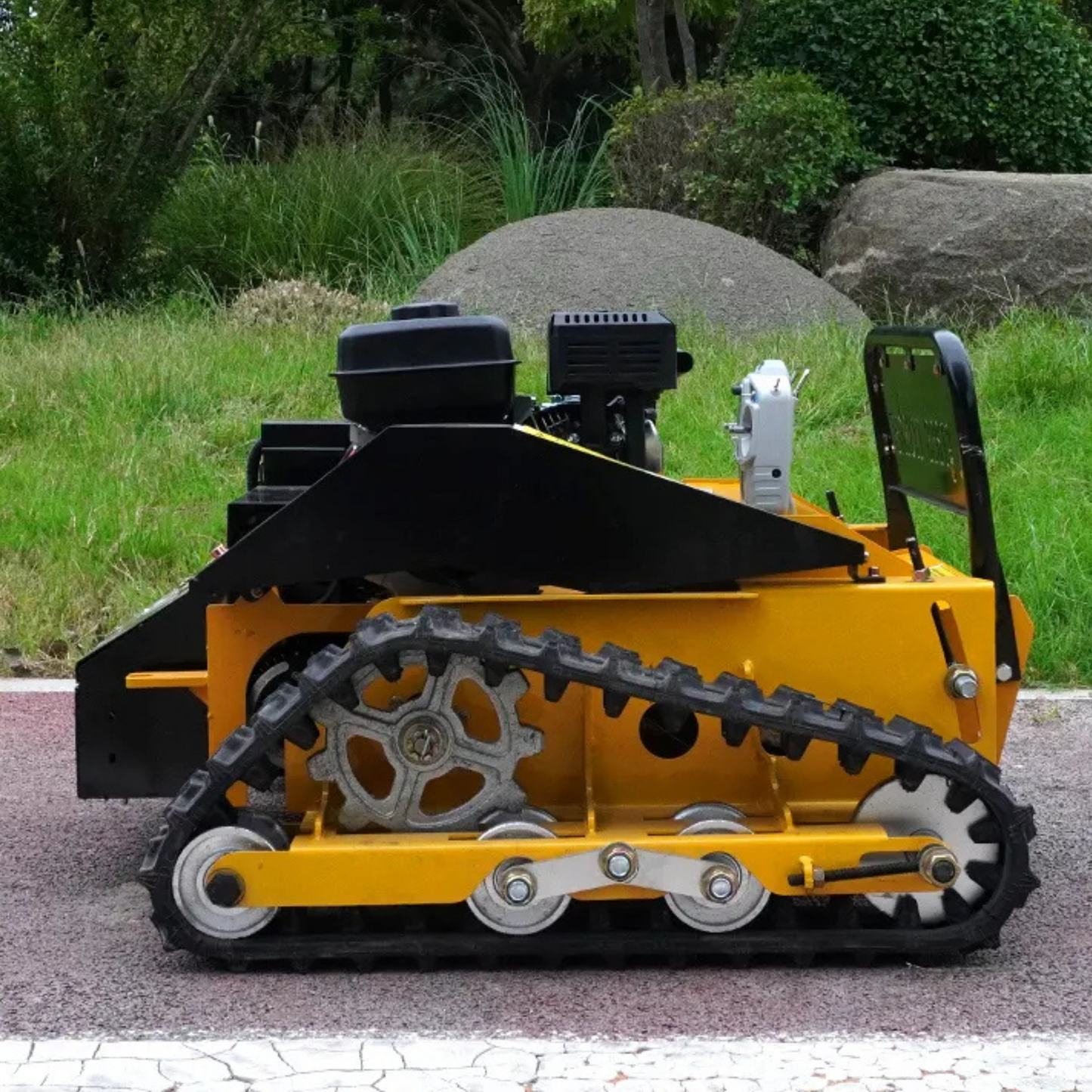 Yellow tracked RC lawn mower on a paved surface with grass and rocks in the background