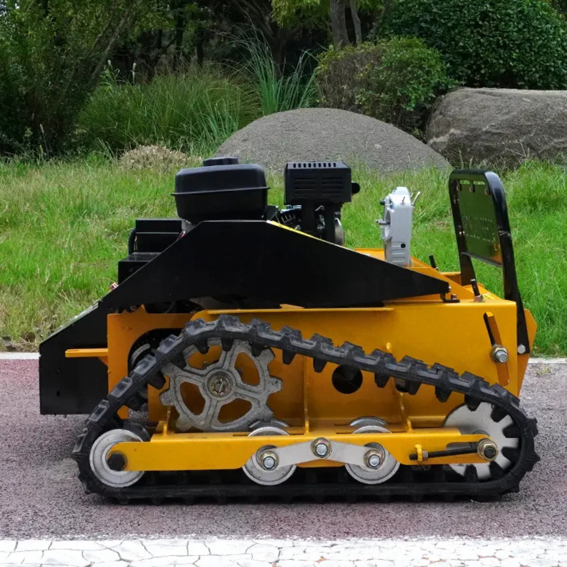 Yellow tracked RC lawn mower on a paved surface with grass and rocks in the background